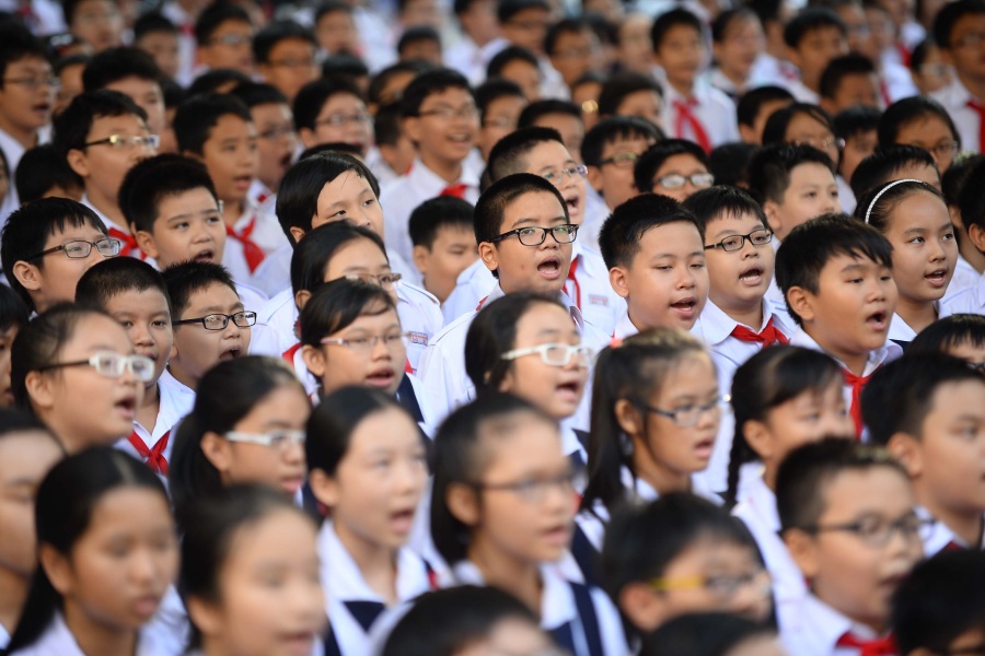 A large group of Vietnamese schoolchildren in uniform sing or recite together during a school assembly; many of them are wearing glasses, highlighting a high prevalence of myopia.